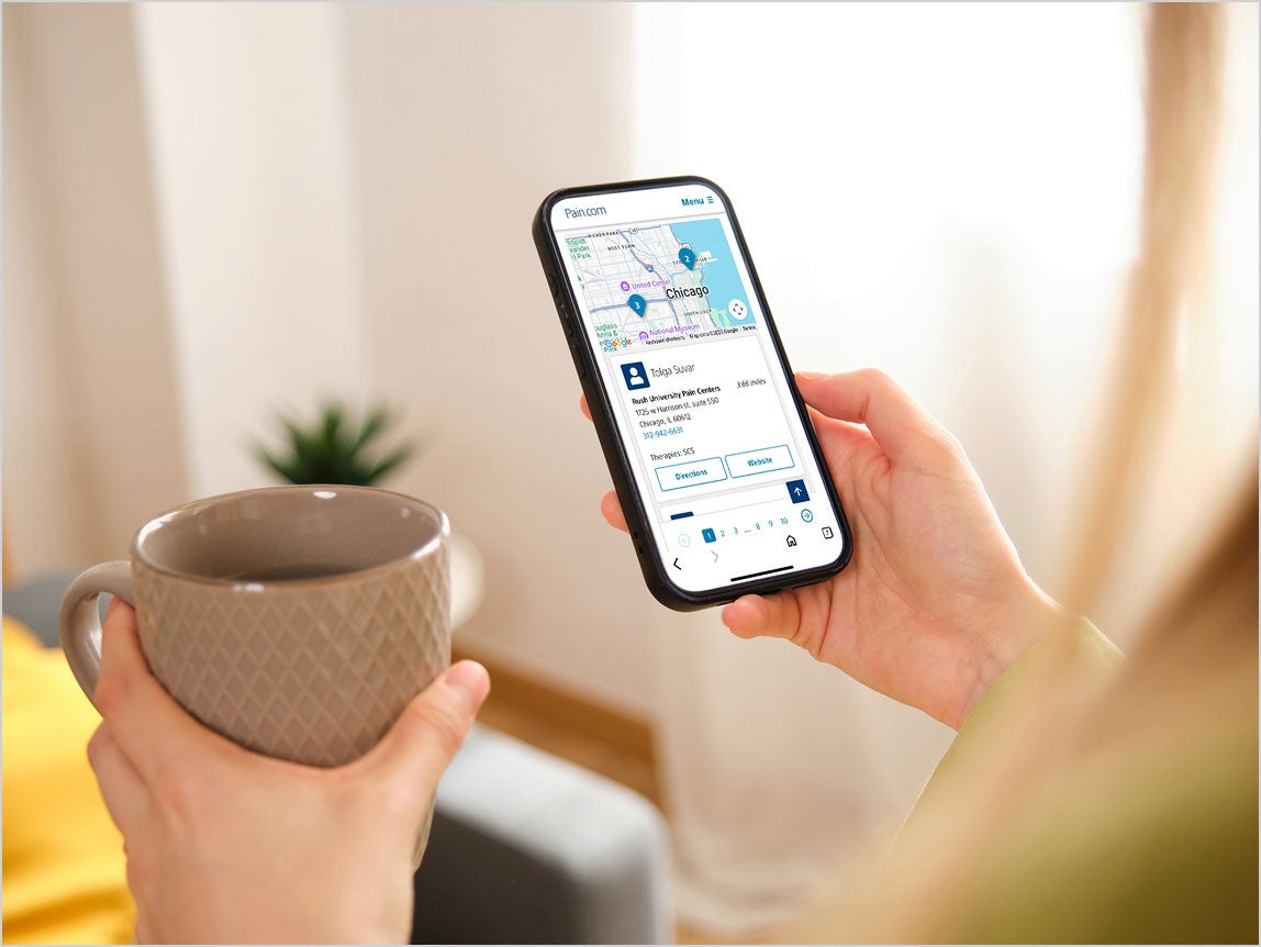 A person holds a smartphone displaying the physician locator in one hand and a mug in the other.
