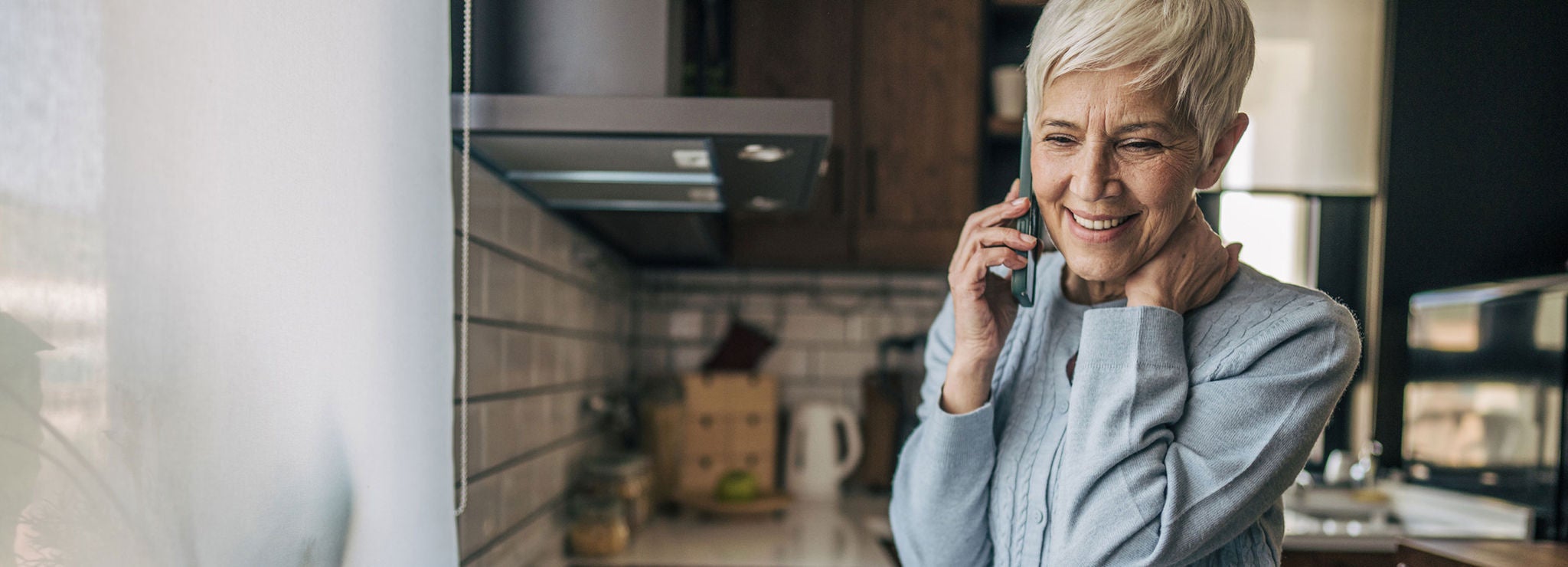 A smiling person with short hair talks on the phone in a kitchen.