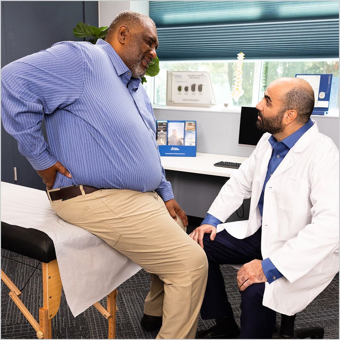A doctor talks with a patient, grimacing and holding their lower back on an exam table.