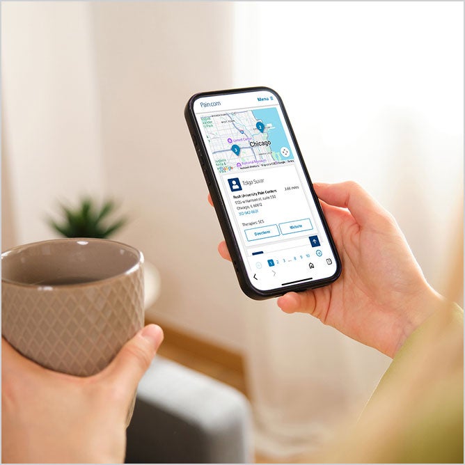 A person holds a smartphone displaying the physician locator in one hand and a mug in the other.