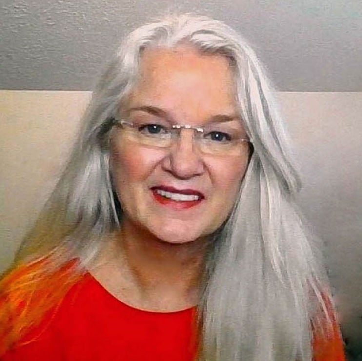 Robin M., smiles. With long gray hair and glasses, she’s wearing a bright red top.
