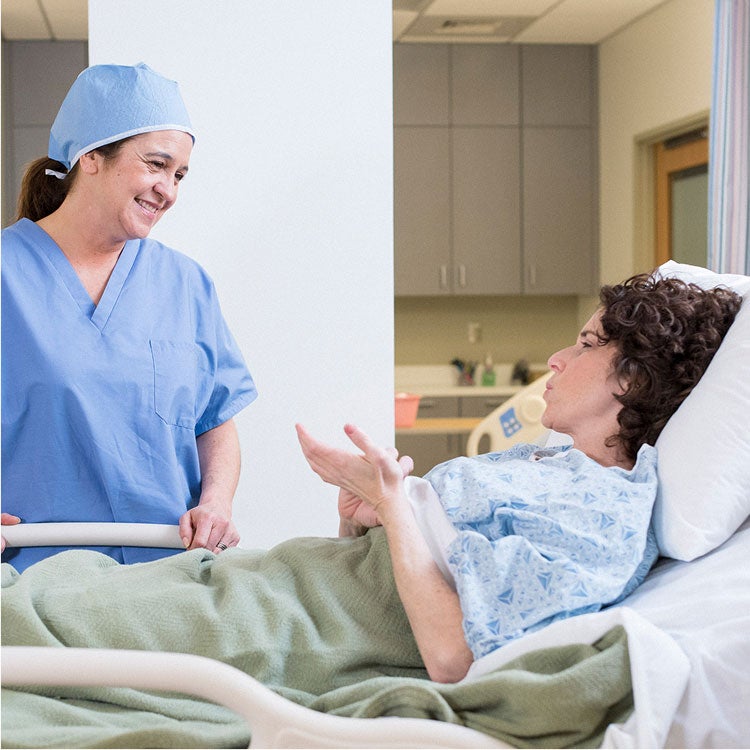 A healthcare provider in scrubs smiles at a patient lying and gesturing while talking in a hospital bed.
