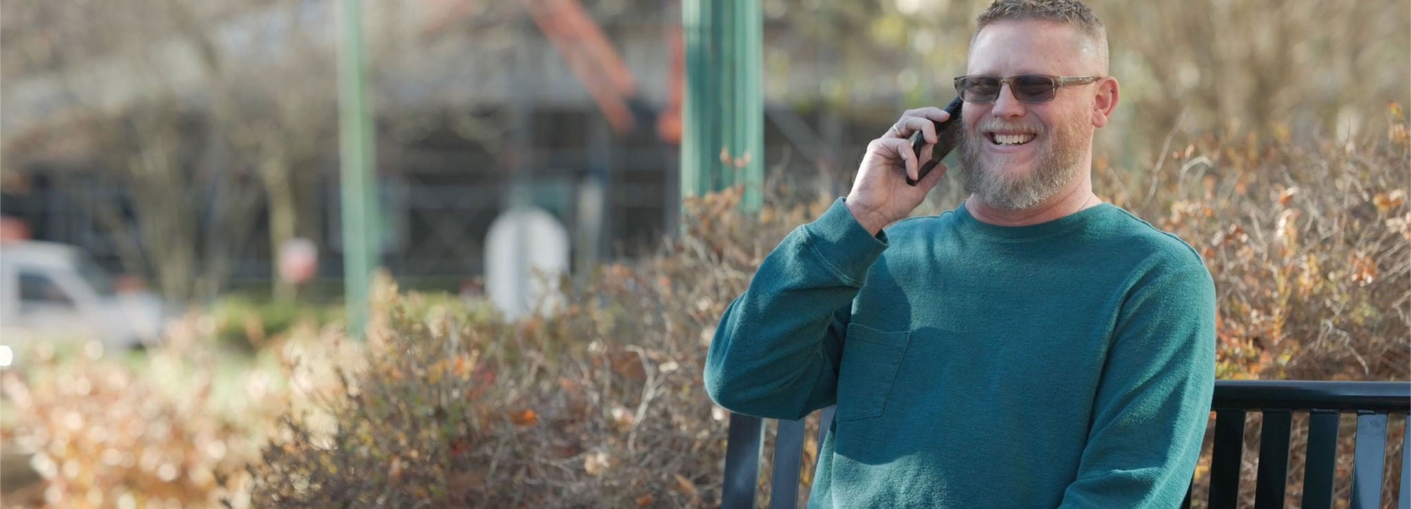 A smiling man with glasses talks on a smartphone while sitting in a park.
