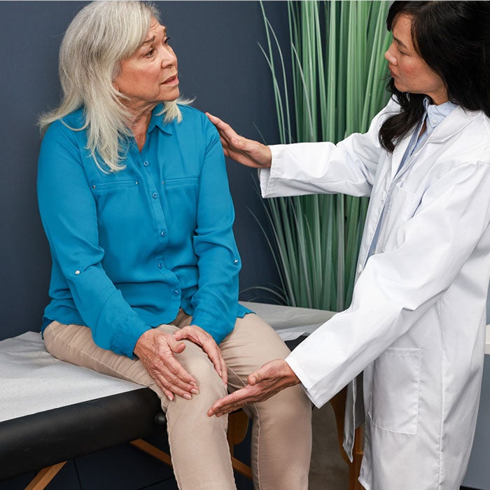 A healthcare provider in a white coat chats with a patient sitting on an exam table holding her knee.