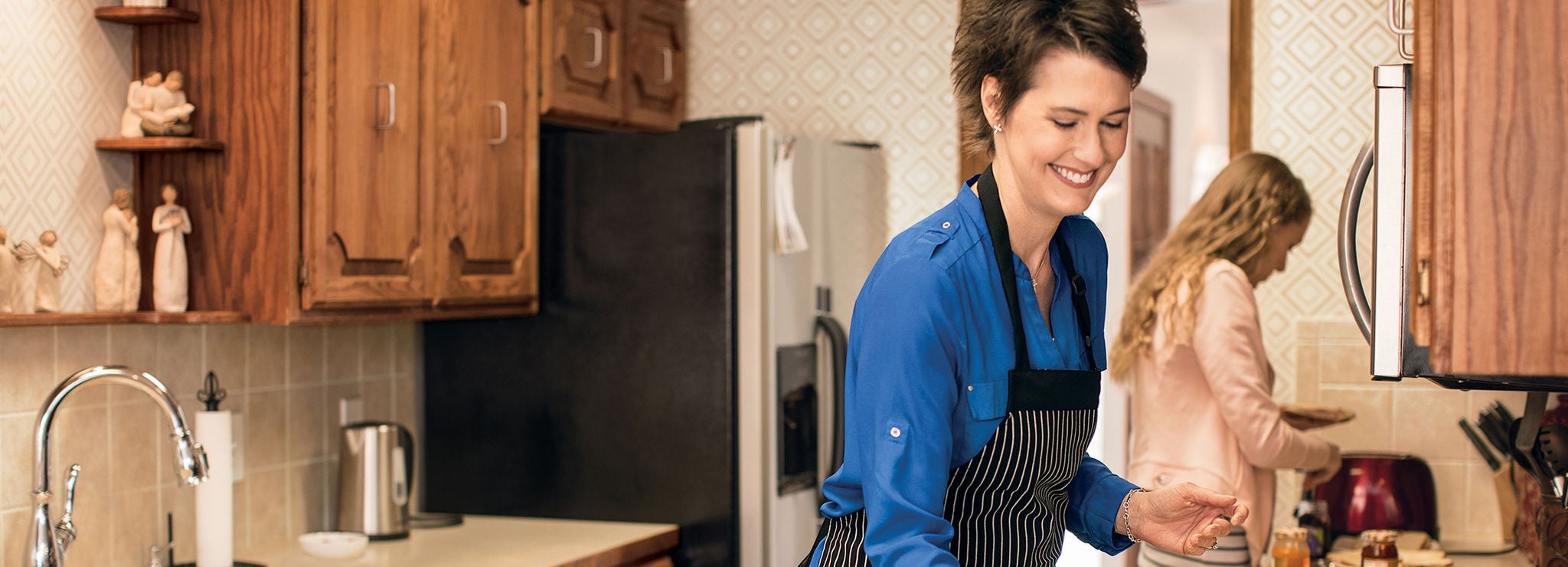 A woman in a black apron smiles while cooking in a kitchen with another woman. 