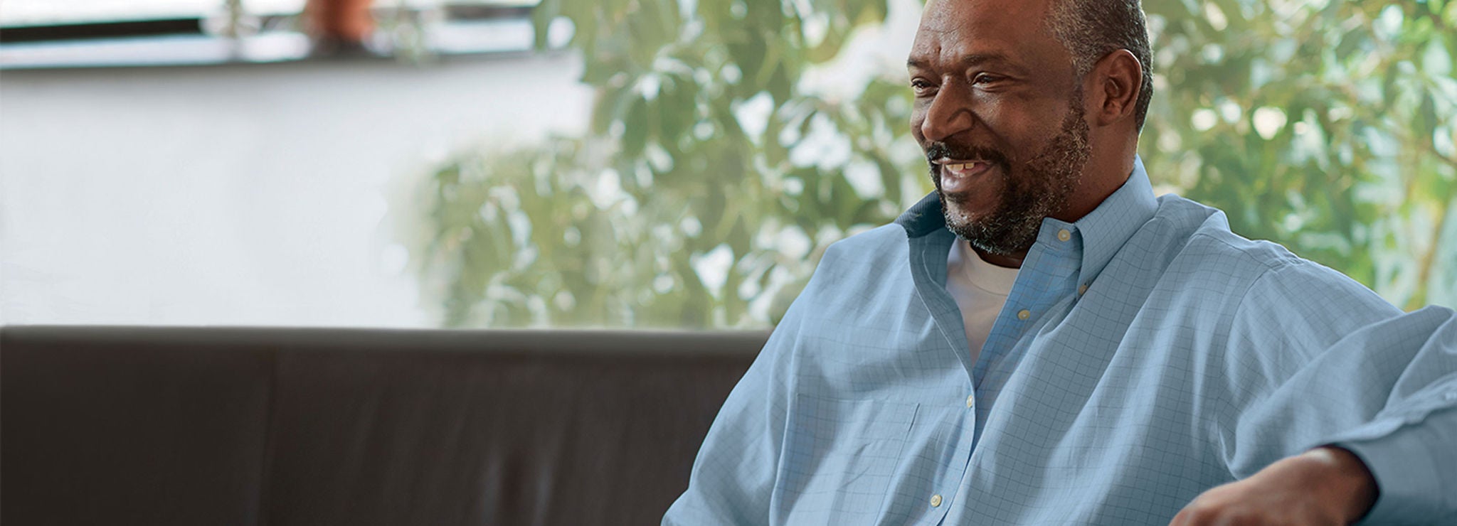 A smiling person sits near a window in front of greenery.