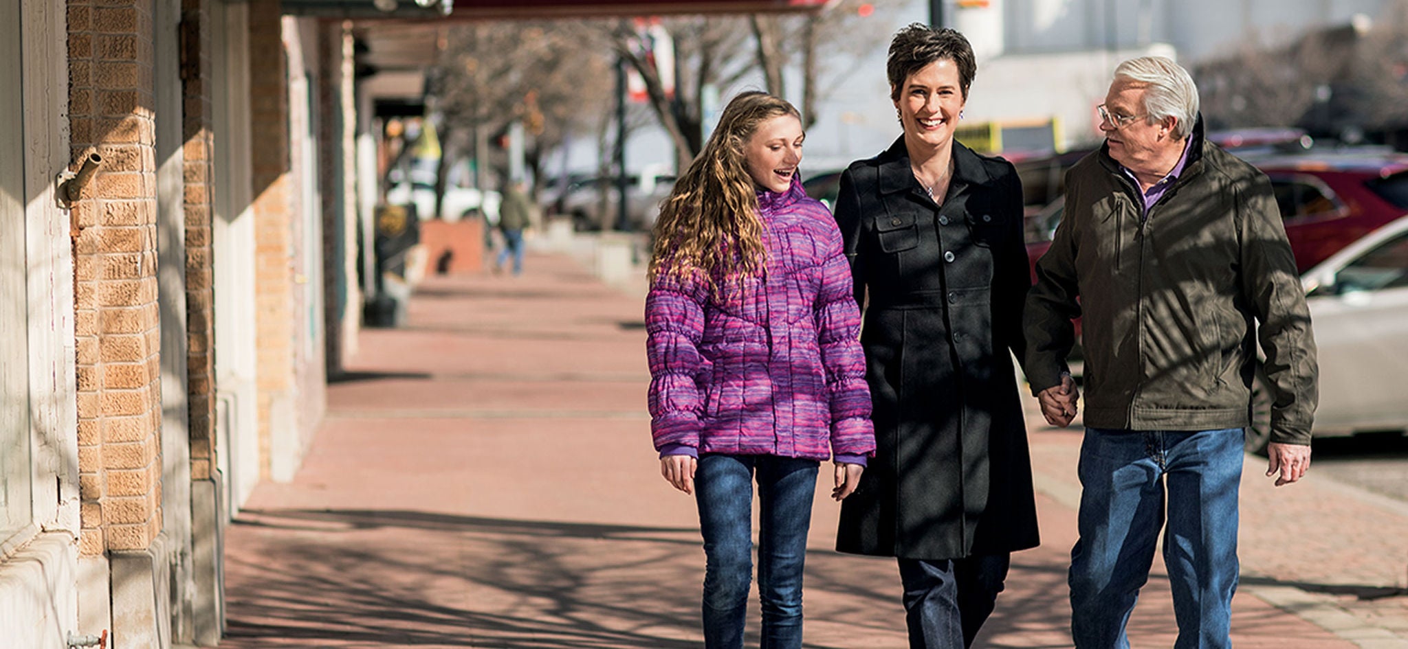 A girl in a purple jacket smiles as she walks and chats with two adults on a sunny sidewalk.