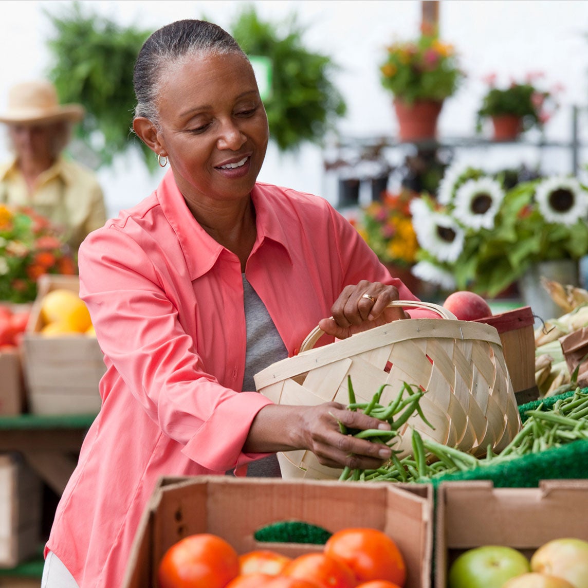A woman in a pink shirt selects green beans from a basket at a farmers market. 