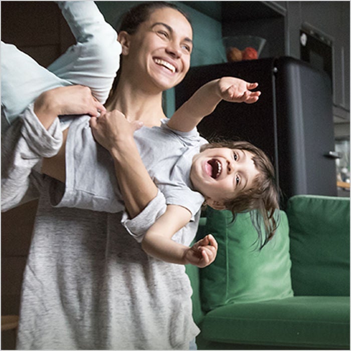 A smiling woman lifts a laughing child sideways in front of a green couch. 