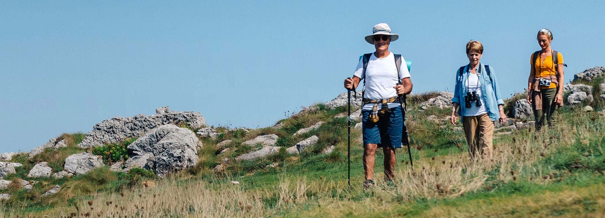Three hikers walk on a grassy path, carrying hiking poles and cameras, with rocks behind them.