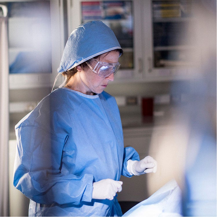 A medical professional in scrubs and goggles focuses on a task.