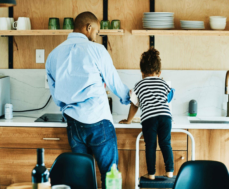 A person stands in front of a kitchen sink with a child standing beside them on a stool.