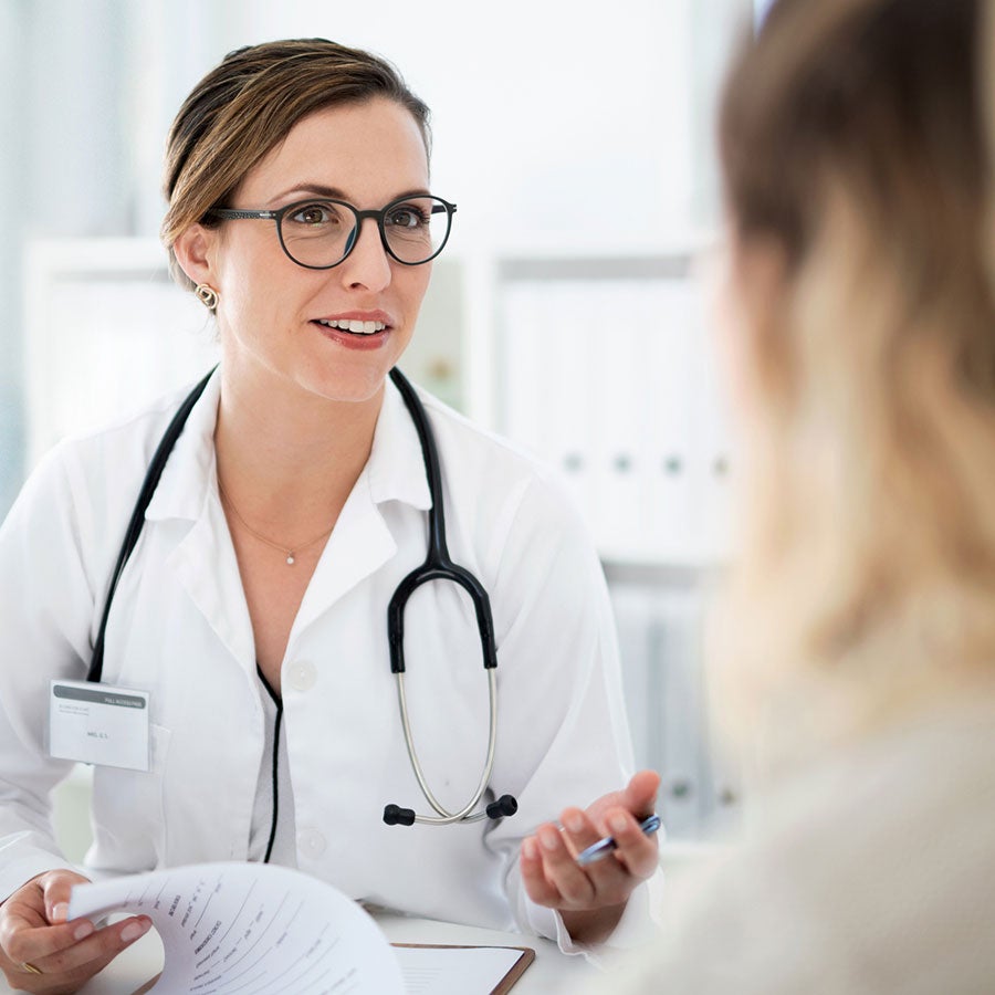 A smiling healthcare provider in a lab coat talks to a patient, holding papers in their hand.