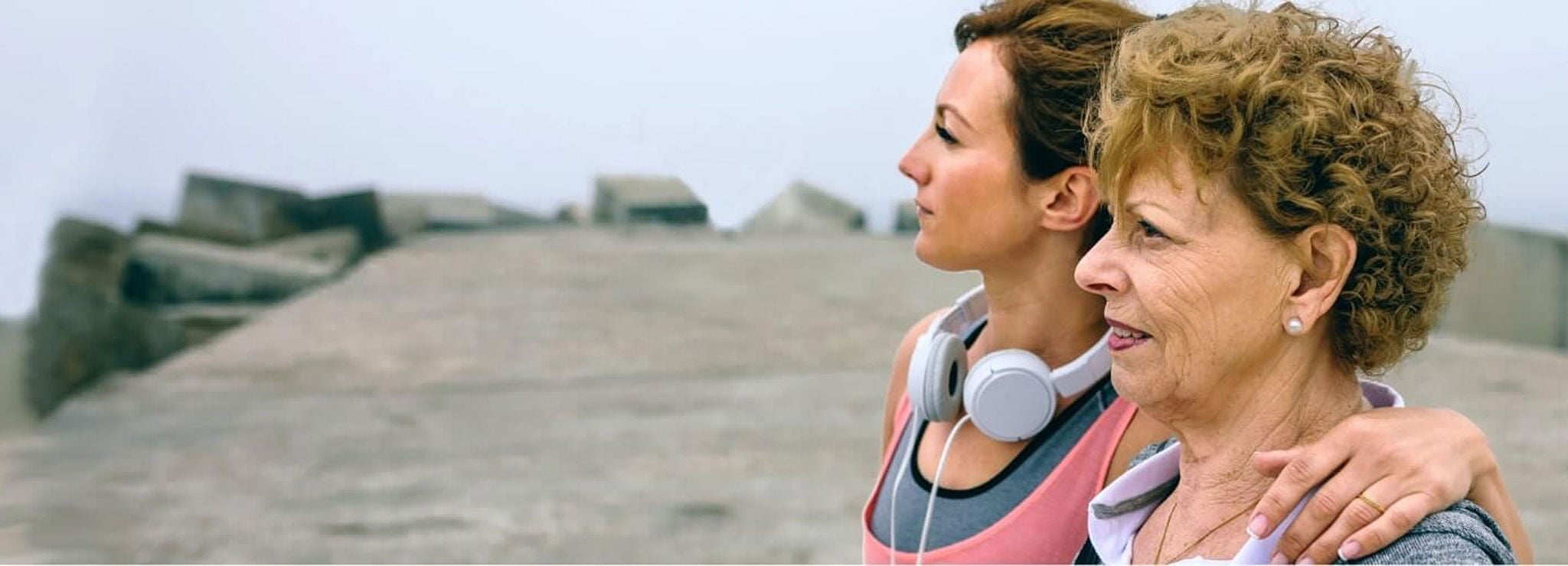 A young person with headphones stands on a beach with their arm around the shoulders of an older person.