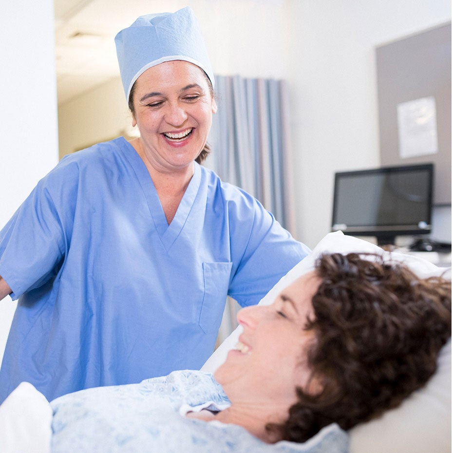 A healthcare provider in scrubs smiles at a patient lying in a hospital bed. 