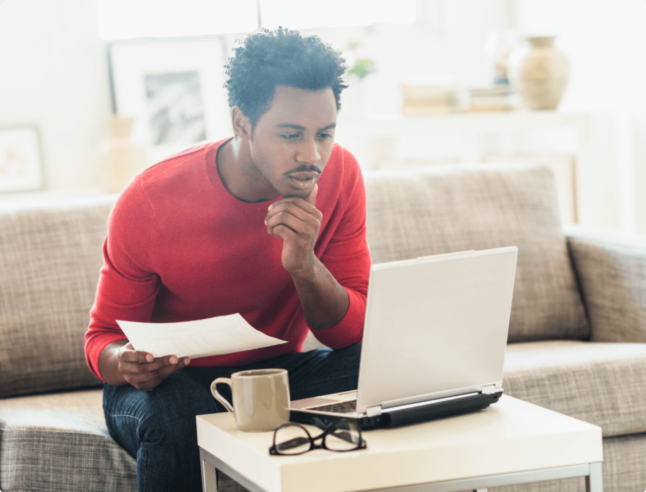 A man holding a document looking at a laptop screen.