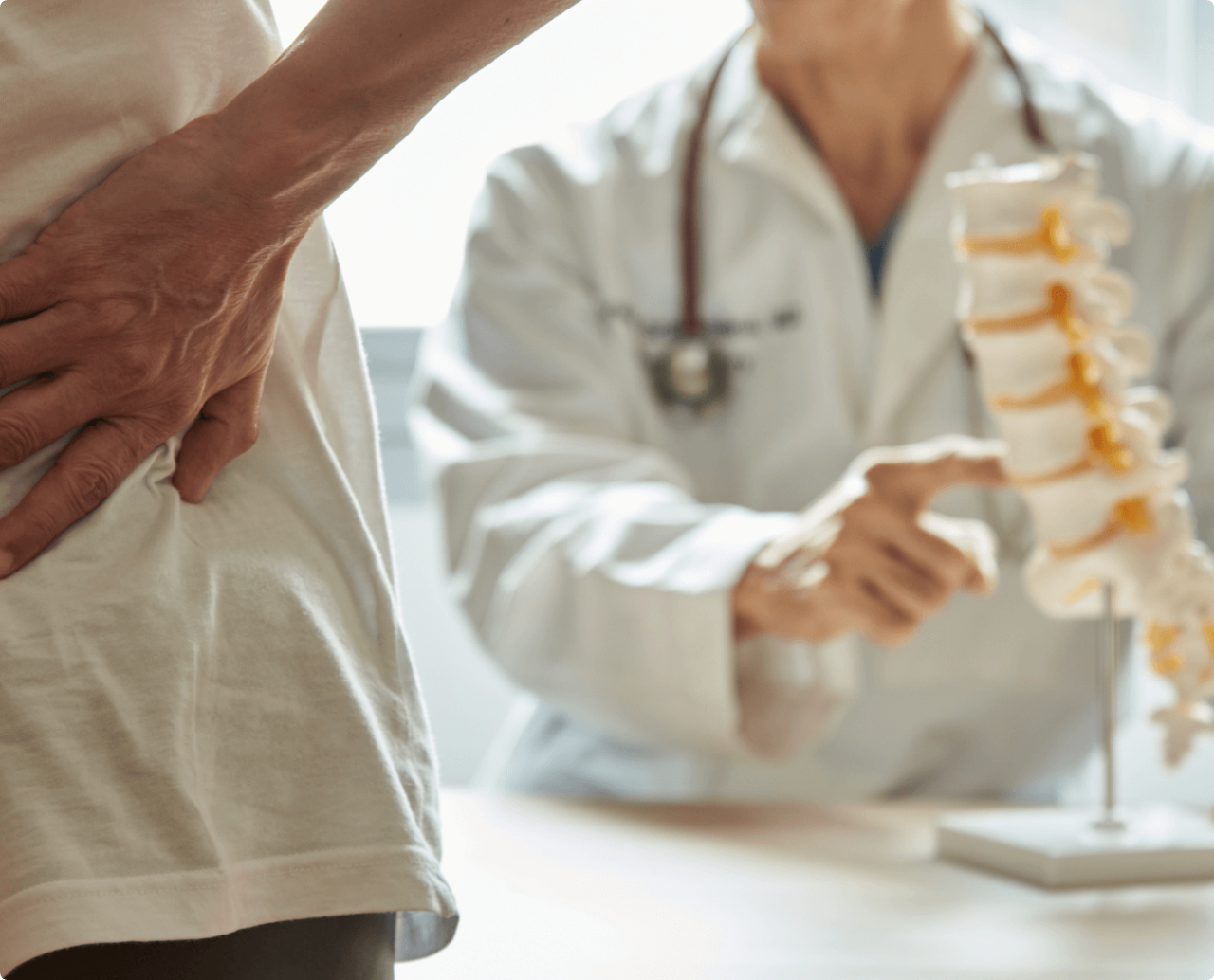 A doctor pointing to a section of the spine while a woman holds her lower back in the foreground.