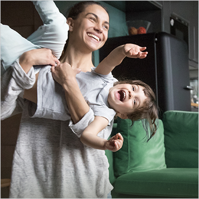 A smiling woman lifts a laughing child sideways in front of a green couch. 