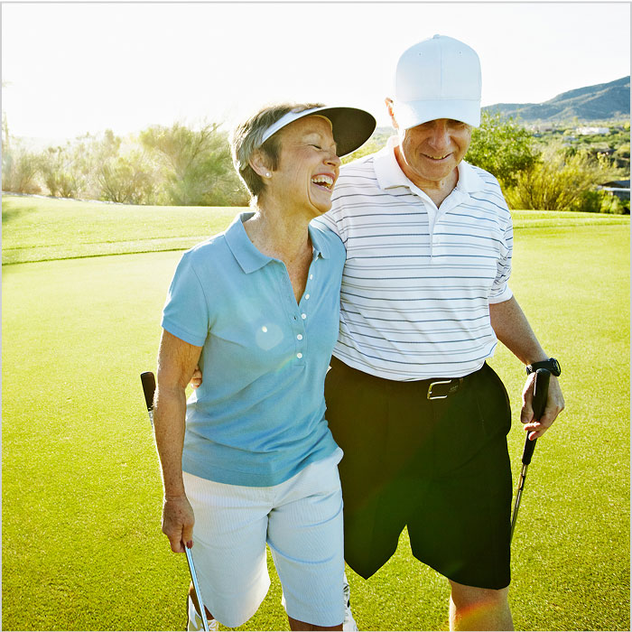 An older couple walks and laughs together on a green, holding golf clubs.