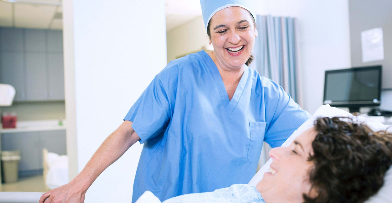 Nurse in scrubs smiling while talking with a patient in hospital bed.