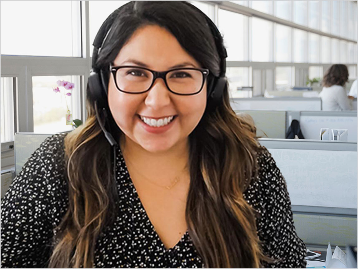 A smiling person with glasses and a headset sits in an office with cubicles.