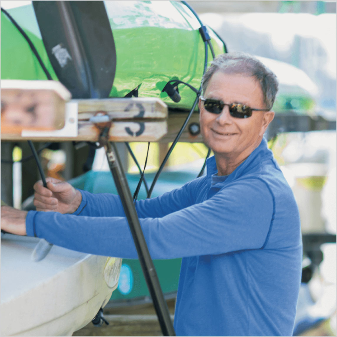 A smiling person in sunglasses adjusts kayaks on a rack outdoors.