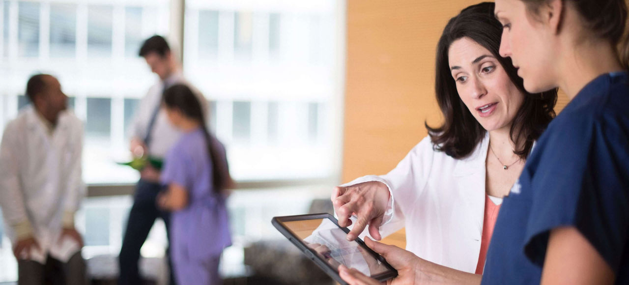 A healthcare professional in a lab coat shows a tablet screen to a nurse.