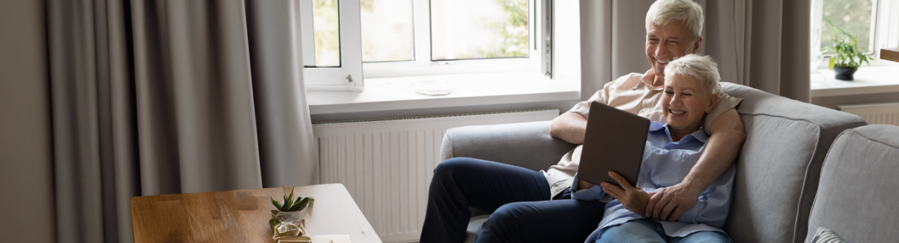 Two older adults sit together on a sofa, smiling while looking at a tablet in a bright living room.