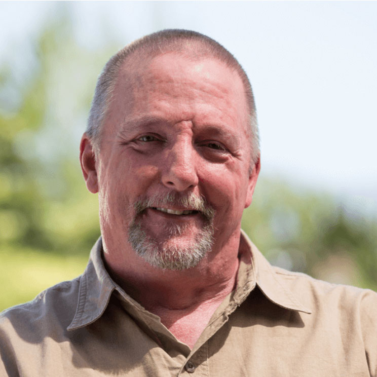 Dana C. smiles. With short gray hair, a beard, and wearing a tan shirt.