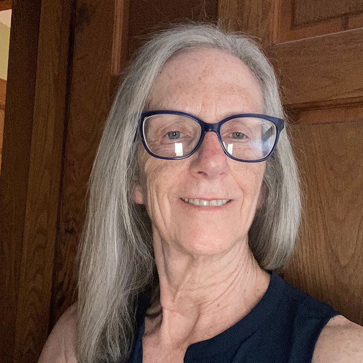 Pam L. smiles. With long gray hair and glasses in front of a wooden background.