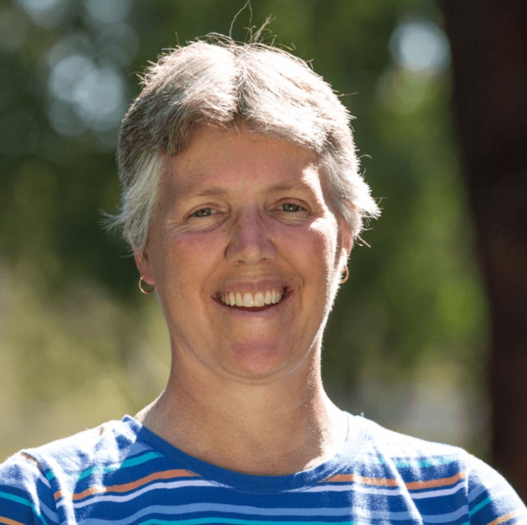 Sandy F. smiles. With short gray hair, she’s in a blue striped shirt.