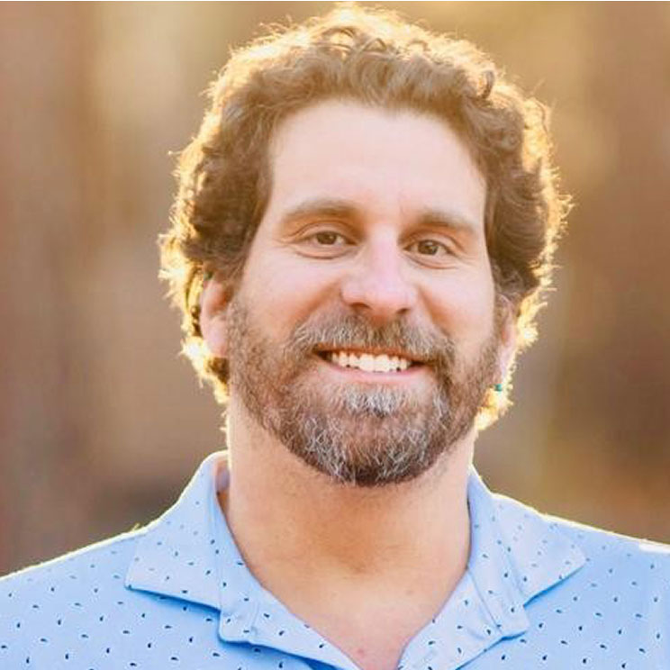 Dave N. smiles. With curly hair and a light blue shirt with small dots