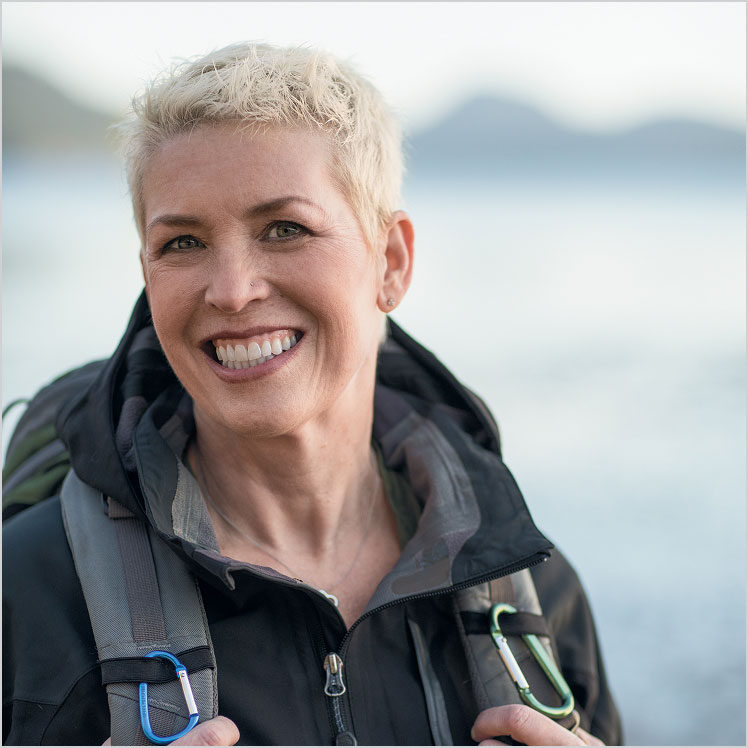 Mary, a smiling person with short hair and outdoor gear, stands near water.