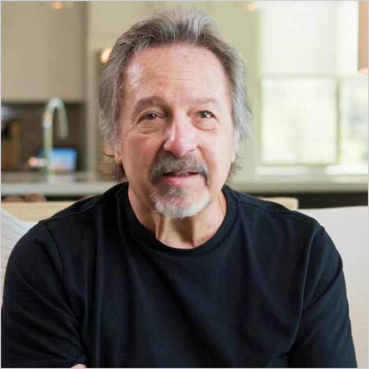 Michael, a person with gray hair and a mustache, smiles while sitting in a kitchen.