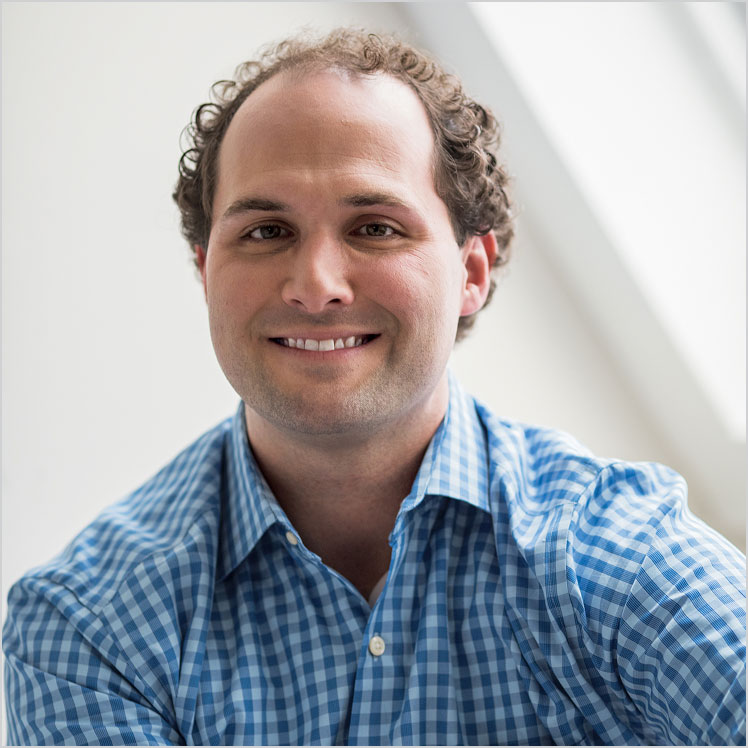 Chris, a smiling person with curly hair and a blue checkered shirt, sits indoors.