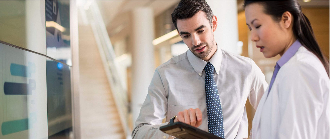 A person in a shirt and tie shows something on a tablet to a doctor in a lab coat.