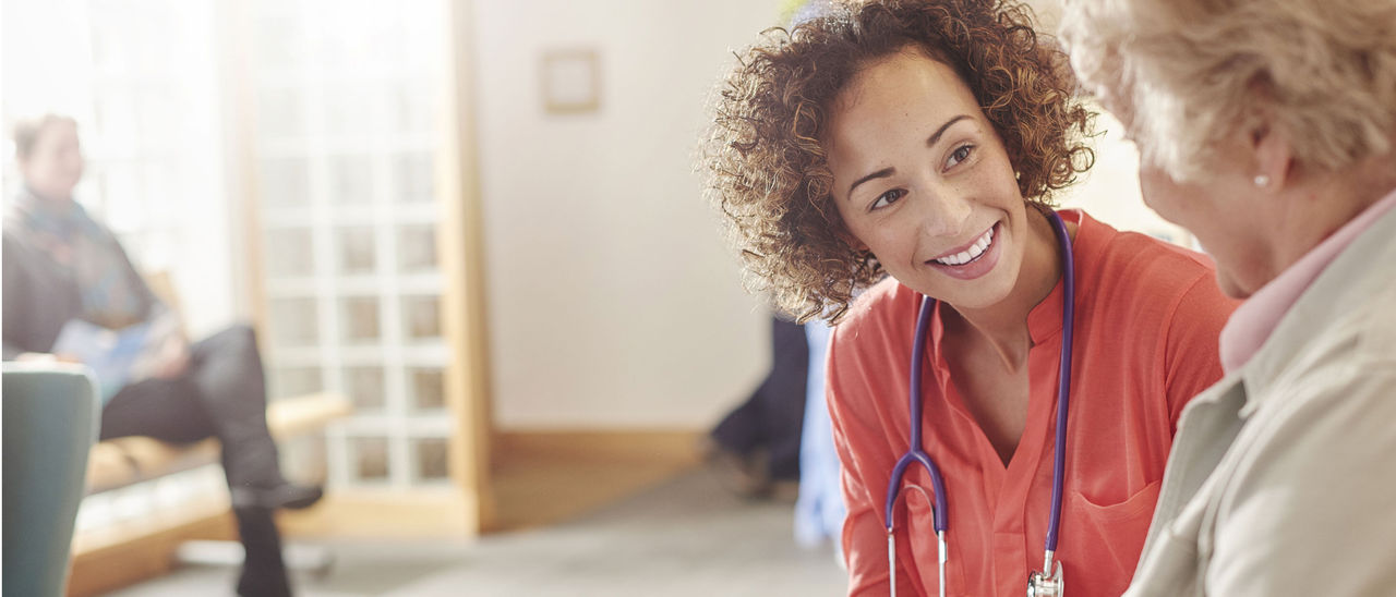 A healthcare provider with curly hair in an orange shirt smiles as she listens to a patient.