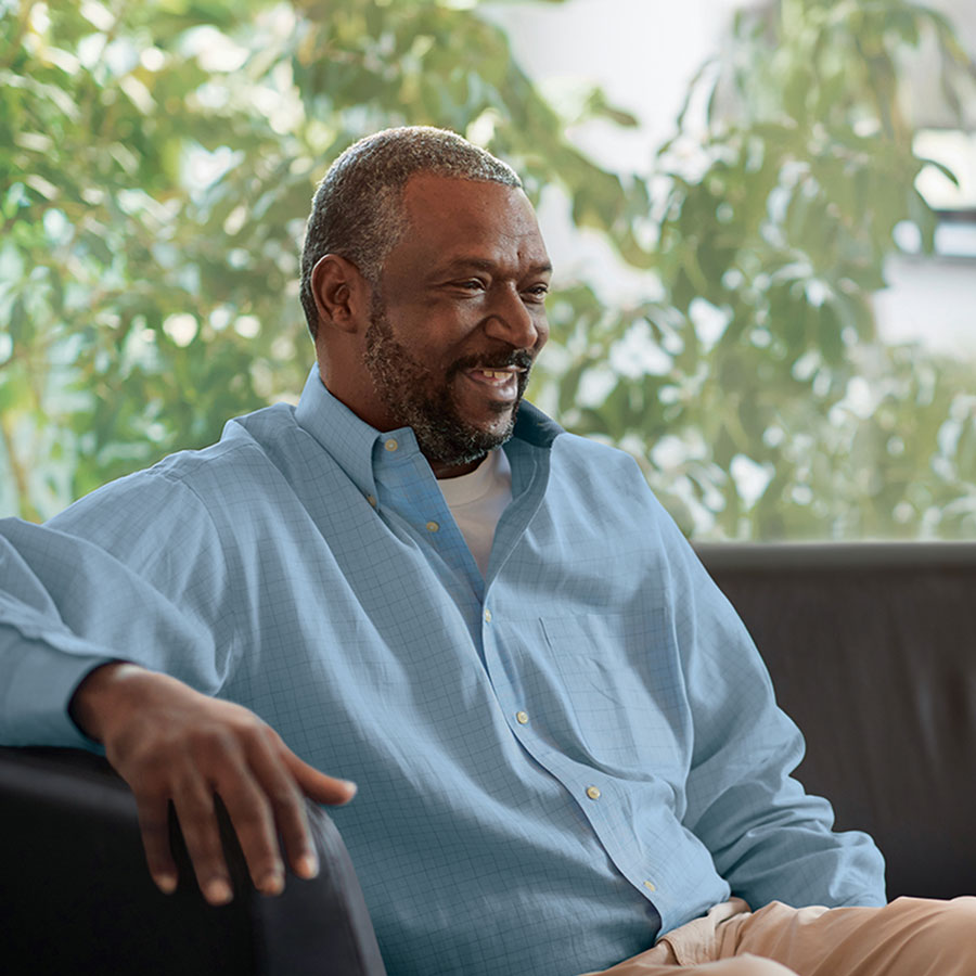 A person with a beard in a light blue shirt sits on a couch with greenery behind them.