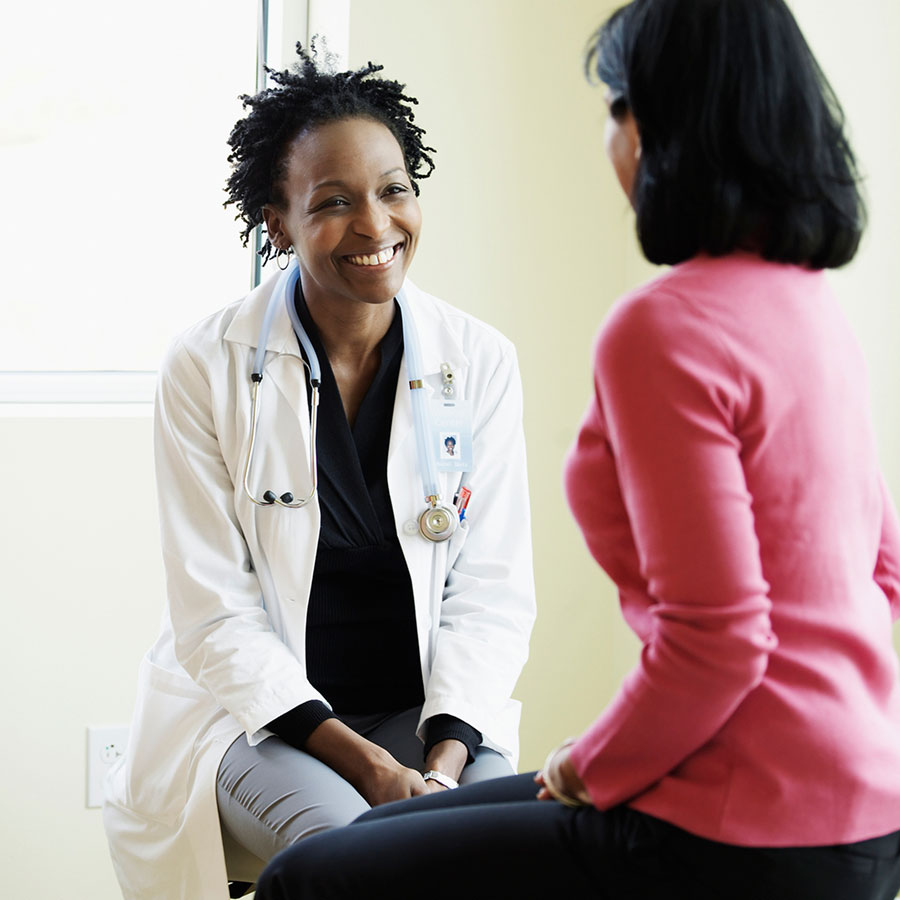 A healthcare provider in a white coat smiles while listening to a patient in a pink sweater.