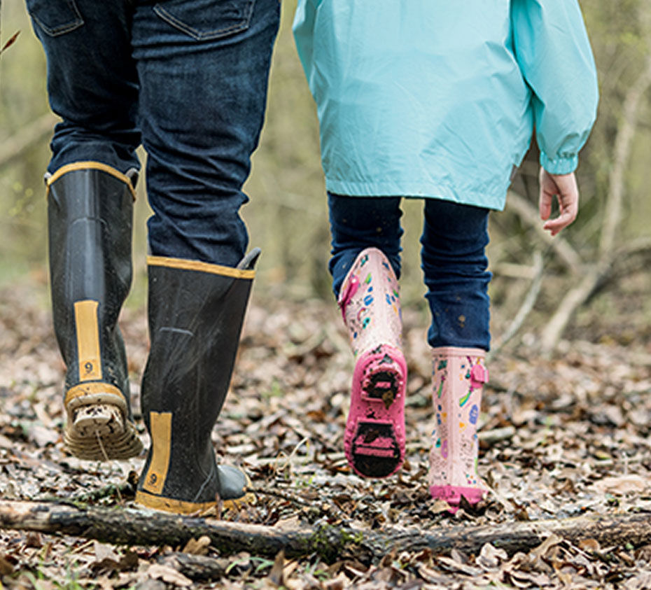 A child and an adult in rain boots walk on a leaf-covered path.