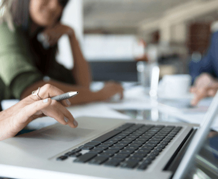 A hand holds a pen while typing on a laptop keyboard during a meeting with other people.