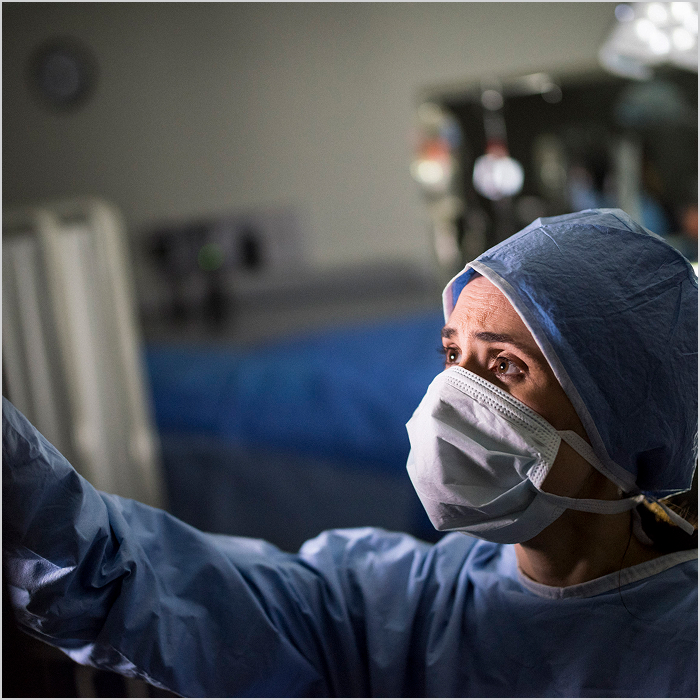 A healthcare worker in a surgical gown and mask looks intently at a medical device.