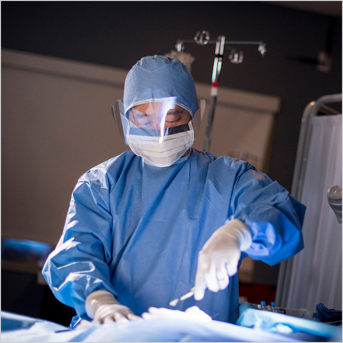 A surgeon wears a face shield and holds a scalpel.